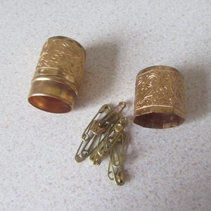 May include: Two gold-coloured metal containers with intricate floral designs. The containers are open and reveal a set of safety pins.