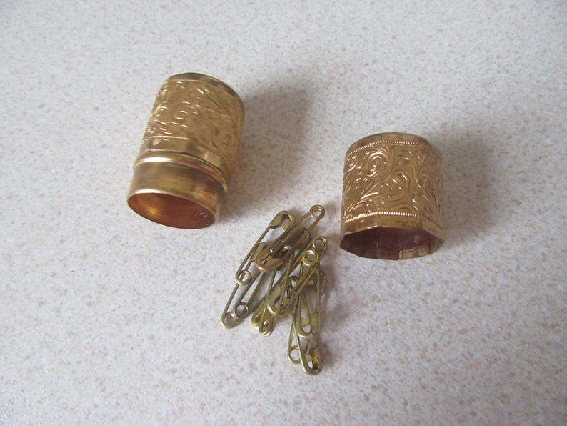 May include: Two gold-coloured metal containers with intricate floral designs. The containers are open and reveal a set of safety pins.