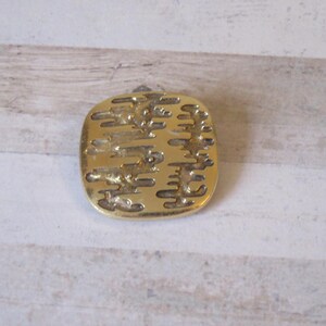 K&ouml;nnte beinhalten: Eine goldfarbene Brosche aus Metall mit einem strukturierten Design aus vertikalen Linien und Wirbeln.