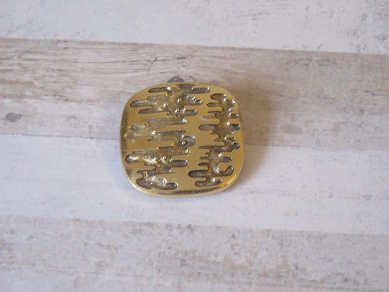 K&ouml;nnte beinhalten: Eine goldfarbene Brosche aus Metall mit einem strukturierten Design aus vertikalen Linien und Wirbeln.