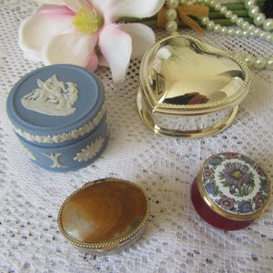 Op de afbeelding: Een assortiment decoratieve sieradendoosjes in verschillende vormen en ontwerpen. Inclusief een blauw-wit keramieken doosje, een goudkleurig hartvormig doosje, een schelpvormig doosje en een doosje met bloemmotief. De doosjes worden gepresenteerd op een wit kanten oppervlak.