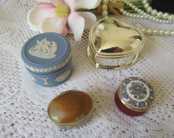 May include: An assortment of decorative trinket boxes in various shapes and designs. Includes a blue and white ceramic box, a gold heart-shaped box, a shell-shaped box, and a floral-patterned box. The boxes are displayed on a white lace surface.