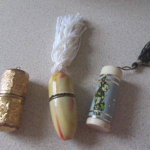 May include: Three vintage thimble cases. One is gold with an ornate design, another is ivory with a white tassel, and the third is blue with a white tassel and a cartoon image of a man in a kilt. The blue thimble case has the text "Bonni Vila" printed on it.
