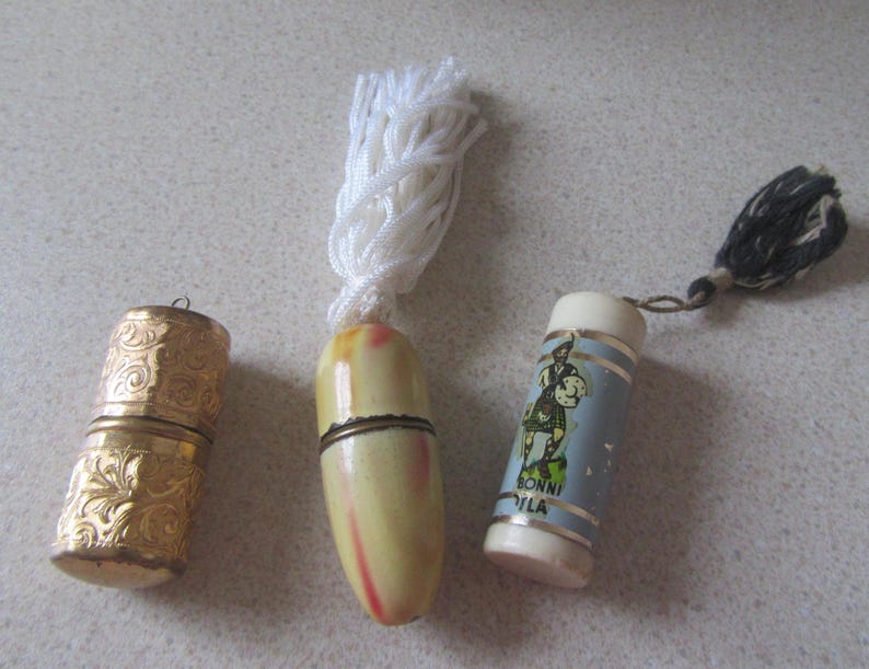 May include: Three vintage thimble cases. One is gold with an ornate design, another is ivory with a white tassel, and the third is blue with a white tassel and a cartoon image of a man in a kilt. The blue thimble case has the text "Bonni Vila" printed on it.