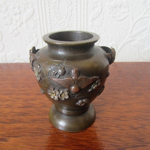 May include: A small, dark bronze vase with intricate floral and bird designs. The vase has two handles and a rounded body.
