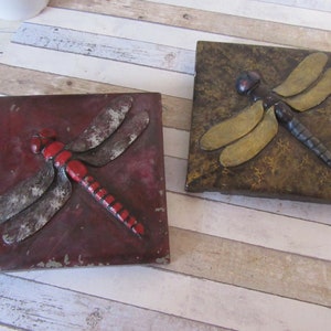 May include: Two square decorative tiles featuring dragonfly designs. One tile is a deep red with a red and silver dragonfly. The other tile is brown with a gold and black dragonfly. Both tiles have a distressed, vintage appearance.