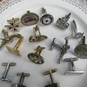 May include: A collection of assorted metal cufflinks featuring various designs including a vintage car, a compass, a lion emblem, a Maltese cross, and coin-style motifs, all displayed together on a white plate with a decorative red and gold rim.