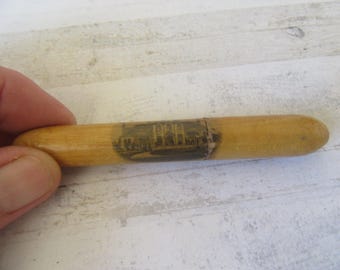May include: A vintage wooden needle case with a cylindrical shape. The case is light brown with a dark band featuring a faded image of a building. The case is held in a hand against a light-colored wooden surface.