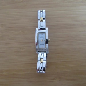 Peut inclure: Montre rectangulaire argentée avec un cadran blanc et des accents dorés. La montre a un bracelet argenté avec des détails dorés. Le cadran de la montre a un petit diamant en haut. La montre est sur une surface en bois.