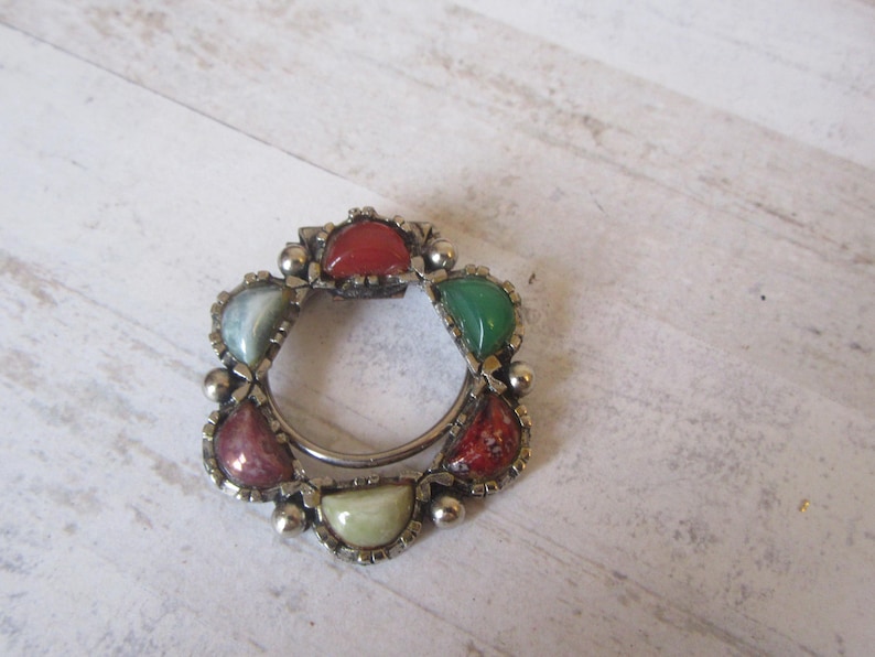 May include: A silver ring with six different colored stones set in the metal. The stones are green, red, and white.