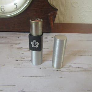 May include: A silver lipstick tube with a black band featuring a white floral design. The cap is detached and placed next to the lipstick. The lipstick is open, revealing the red lipstick inside. A wooden clock is in the background.