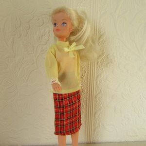 May include: A vintage doll with blonde hair and blue eyes, dressed in a yellow top with a bow and a red plaid skirt. The doll's outfit includes white lace cuffs. The doll is positioned against a textured wall.