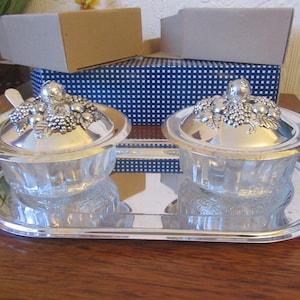 May include: A silver-plated condiment set with two glass bowls, each with a lid adorned with a grape cluster design. The set includes two small spoons and sits on a matching tray. The background includes a blue and white box.