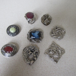 May include: Assortment of eight silver-tone brooches with various coloured stones. The brooches feature a variety of designs, including a Celtic knot, a heart, and a flower.