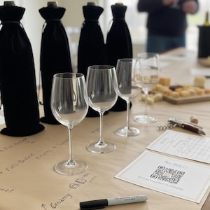 Puede incluir: Una configuración de cata de vinos con copas de vino transparentes, botellas de vino cubiertas y una tabla de quesos. Una cubierta de mesa de papel marrón tiene notas manuscritas y una ilustración de queso. Un marcador negro y una tarjeta de código QR también están sobre la mesa.