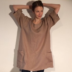 May include: A woman wearing a loose-fitting, light brown linen dress with pockets. She is standing with her arms raised above her head, and her feet are bare.