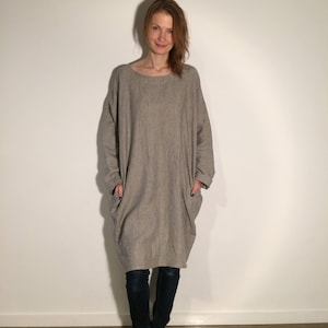 May include: A long, loose-fitting, gray linen dress with long sleeves and large pockets. The dress is worn with dark blue jeans and the model is barefoot.