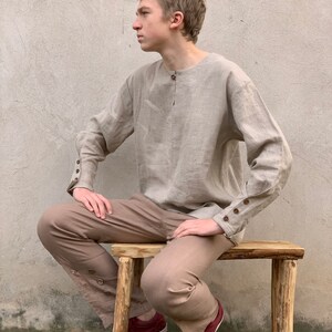 May include: A person wearing a light brown linen shirt with long sleeves and brown pants. The shirt has a button closure at the neck and buttons on the cuffs. The person is sitting on a wooden stool.