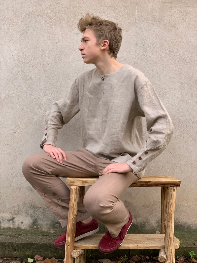 May include: A person wearing a light brown linen shirt with long sleeves and brown pants. The shirt has a button closure at the neck and buttons on the cuffs. The person is sitting on a wooden stool.