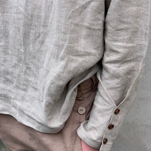May include: A light gray linen shirt with long sleeves and wooden button closures on the cuffs. The shirt is tucked into brown linen pants.