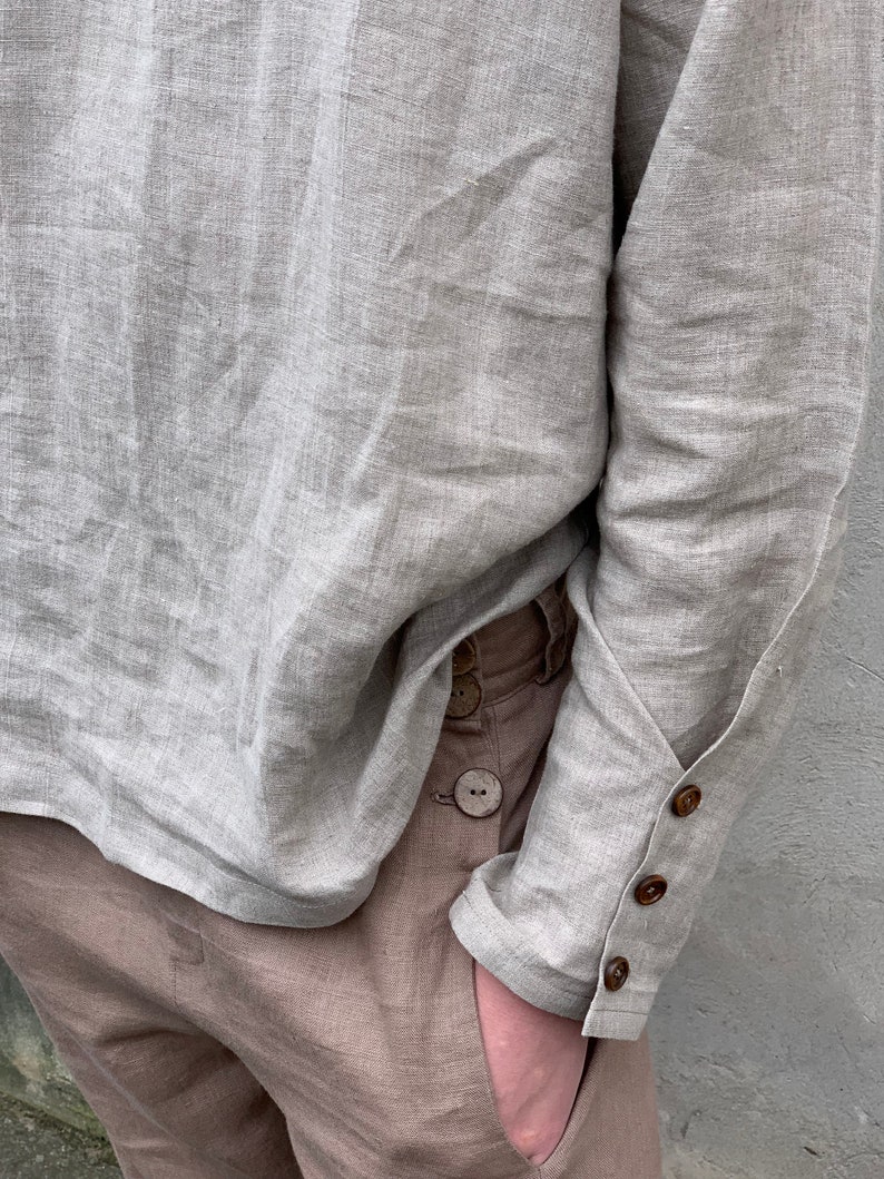 May include: A light gray linen shirt with long sleeves and wooden button closures on the cuffs. The shirt is tucked into brown linen pants.