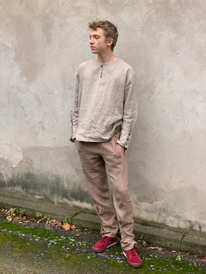 May include: A person wearing a light brown linen shirt with a button-up collar and long sleeves. They are also wearing light brown linen pants with button details on the cuffs. The person is wearing red sneakers.