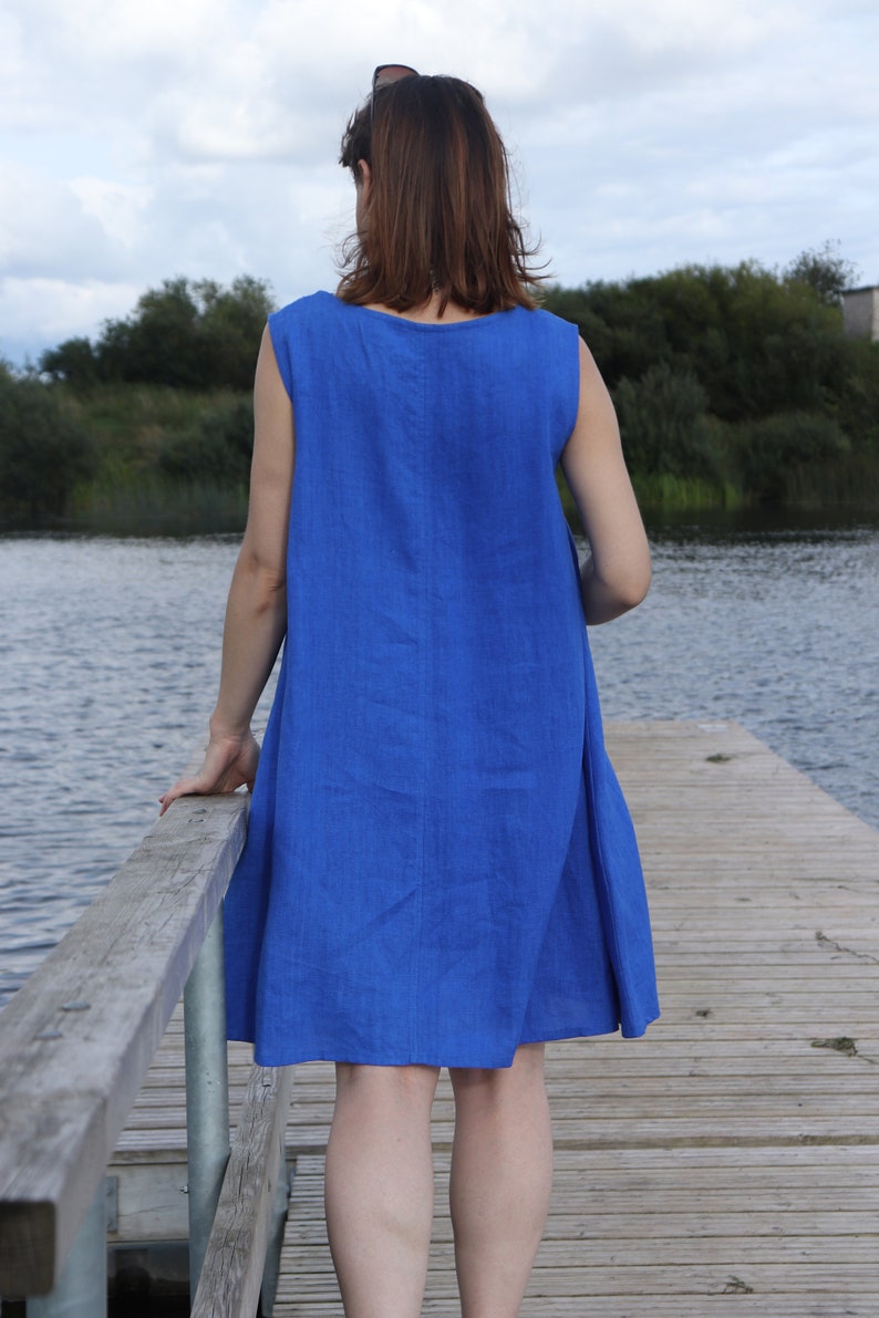Puede incluir: Una mujer con un vestido de lino azul real sin mangas se encuentra de pie en un muelle de madera con vistas a un lago.