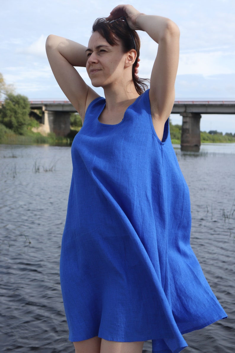 Puede incluir: Una mujer con un vestido de lino azul real sin mangas se encuentra junto a una masa de agua. El vestido tiene un corte sencillo y holgado y un escote redondo.