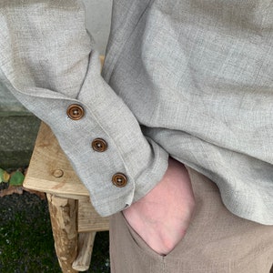 May include: A beige linen shirt with long sleeves and brown button detailing on the cuffs. The shirt is tucked into beige linen pants.