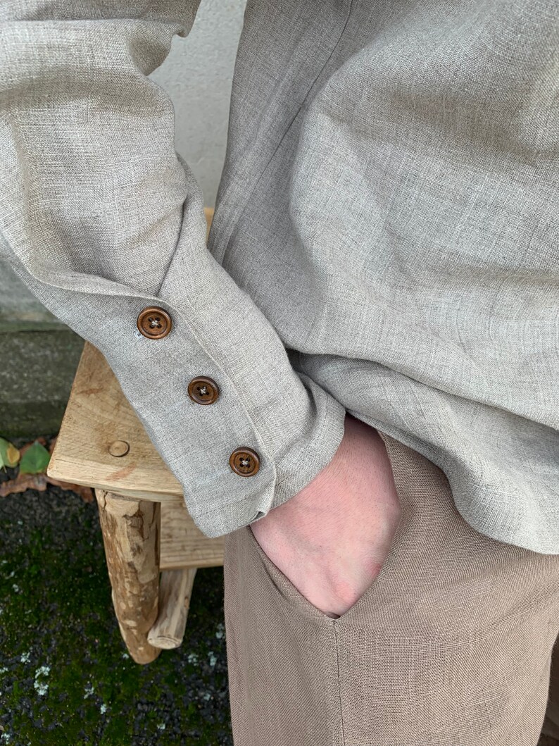 May include: A beige linen shirt with long sleeves and brown button detailing on the cuffs. The shirt is tucked into beige linen pants.