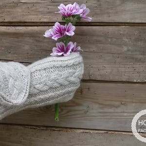 May include: A pair of gray knitted mittens with a cable knit pattern, held by a person, with a bouquet of pink flowers in the foreground.