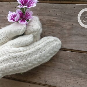 May include: Cream-colored knitted mittens with a cable knit design, holding purple and white flowers. The mittens are displayed against a rustic wooden background. The Home Factory logo is visible in the upper right corner.
