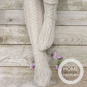 May include: A pair of beige knitted knee-high socks with a cable knit pattern. The socks are worn by a person sitting on a wooden floor. The image includes the text "HOME FACTORY".