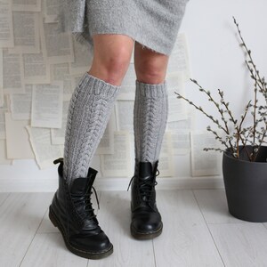 Puede incluir: Un par de calcetines grises de punto hasta la rodilla con un patrón de trenza, usados con botas de cuero negro. Los calcetines se combinan con una falda gris y la persona está de pie frente a una pared cubierta de páginas de libros.