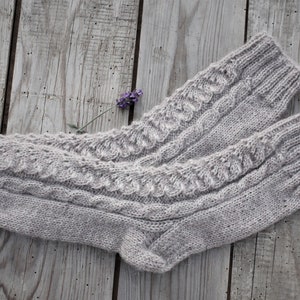Puede incluir: Un par de calcetines de punto grises con un patr&oacute;n de trenza. Los calcetines est&aacute;n sobre una superficie de madera con una peque&ntilde;a flor morada.