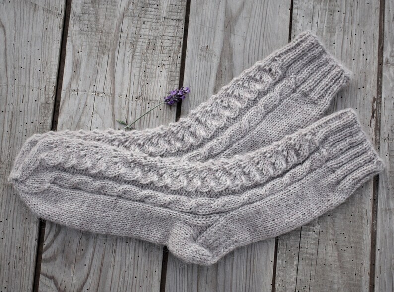 Puede incluir: Un par de calcetines de punto grises con un patr&oacute;n de trenza. Los calcetines est&aacute;n sobre una superficie de madera con una peque&ntilde;a flor morada.