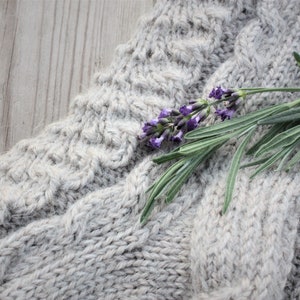 Puede incluir: Una rama de lavanda con flores moradas descansa sobre una tela de punto gris. La tela tiene un patr&oacute;n de punto de cable texturizado.