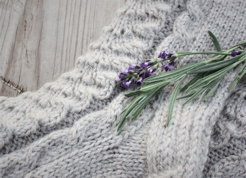 Puede incluir: Una rama de lavanda con flores moradas descansa sobre una tela de punto gris. La tela tiene un patr&oacute;n de punto de cable texturizado.