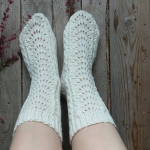 Puede incluir: Un par de calcetines blancos de punto con un patr&oacute;n de encaje. Los calcetines est&aacute;n puestos en los pies de una persona y descansan sobre un suelo de madera.