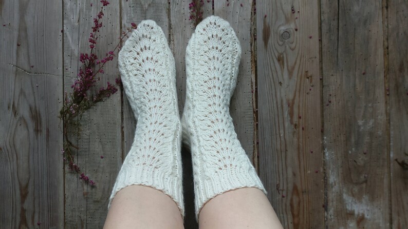 Puede incluir: Un par de calcetines blancos de punto con un patr&oacute;n de encaje. Los calcetines est&aacute;n puestos en los pies de una persona y descansan sobre un suelo de madera.