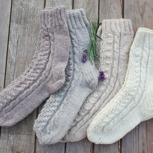 Puede incluir: Cuatro pares de calcetines de punto en varios tonos de beige y blanco. Los calcetines tienen un patr&oacute;n de punto de cable y se muestran sobre una superficie de madera.