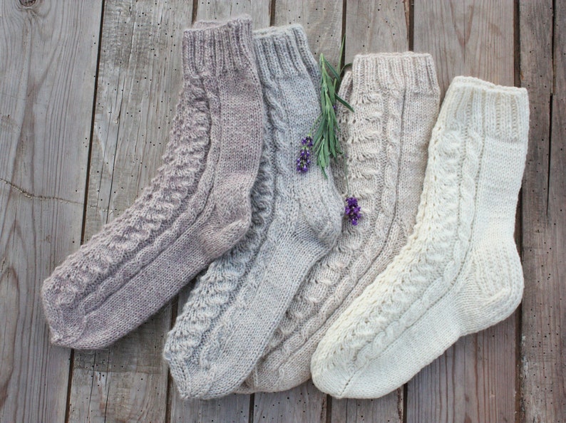 Puede incluir: Cuatro pares de calcetines de punto en varios tonos de beige y blanco. Los calcetines tienen un patr&oacute;n de punto de cable y se muestran sobre una superficie de madera.
