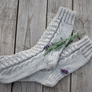 Puede incluir: Un par de calcetines de punto gris con un patr&oacute;n de trenza. Los calcetines est&aacute;n colocados sobre una superficie de madera con una ramita de flores de lavanda.