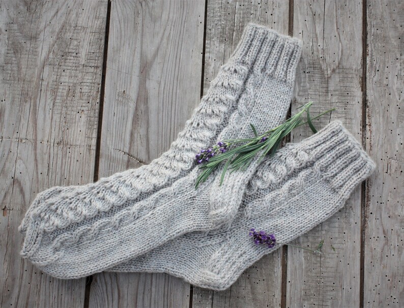 Puede incluir: Un par de calcetines de punto gris con un patr&oacute;n de trenza. Los calcetines est&aacute;n colocados sobre una superficie de madera con una ramita de flores de lavanda.