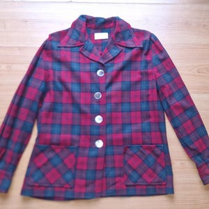 May include: A vintage plaid jacket in shades of burgundy, teal, and navy blue. The jacket features a collared neckline, long sleeves, two front pockets, and is fastened with four white buttons. The fabric has a classic plaid pattern.