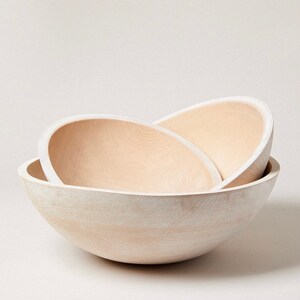 May include: Three wooden bowls of varying sizes, nested together. The bowls are a light beige color and have a smooth, rounded shape.