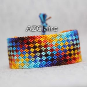 May include: A vibrant woven bracelet featuring a geometric design in blue, orange, yellow, and brown. It has a blue string closure. The text "AZClaire" is visible on the bracelet.