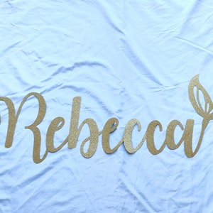May include: Gold glitter name banner with the name "Rebecca" and a mermaid tail cutout. The banner is on a light blue fabric background.