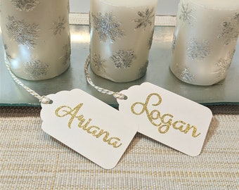 May include: Two white candles with silver snowflake designs and gold glitter names "Ariana" and "Logan" on white gift tags.