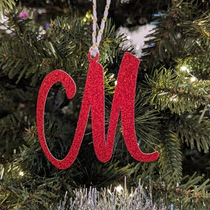 May include: Red glitter letter M Christmas ornament hanging on a green Christmas tree.